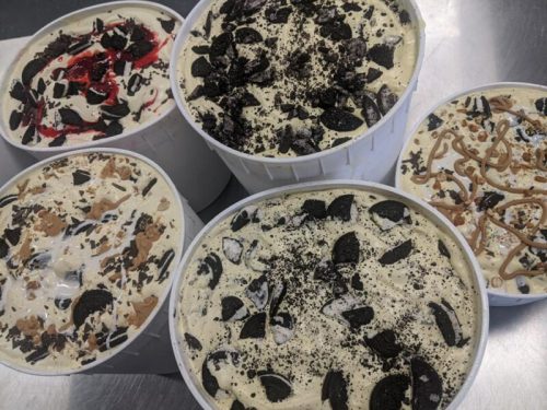 Five large containers of ice cream with various toppings, including crushed cookies, chocolate, and caramel swirls.