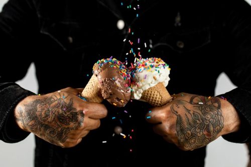A person with tattooed hands holds two ice cream cones, one chocolate and one vanilla, with colorful sprinkles falling.