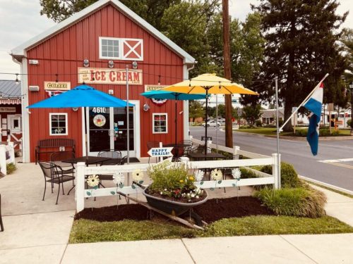 A red ice cream shop with blue and yellow umbrellas, outdoor seating, and a flower garden in front.