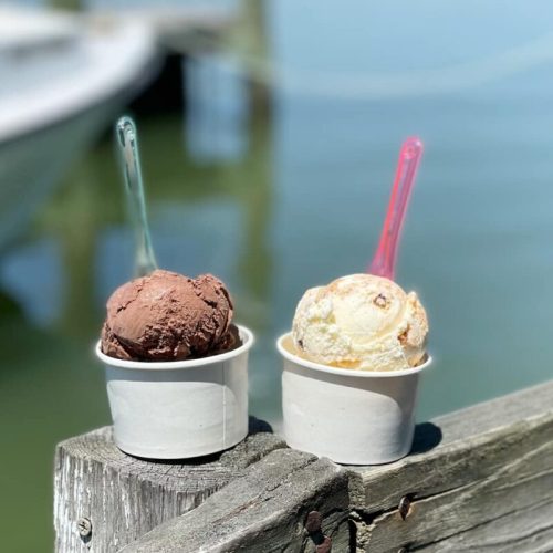 Two cups of ice cream, one chocolate and one vanilla, sit on a wooden post by the water, with a boat in the background.