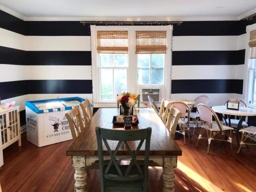 A bright dining area with black and white striped walls, a wooden table, and various chairs, including a box in the corner.