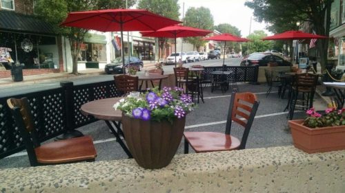 Outdoor dining area with tables, chairs, and red umbrellas, featuring flower pots along a street in a small town.