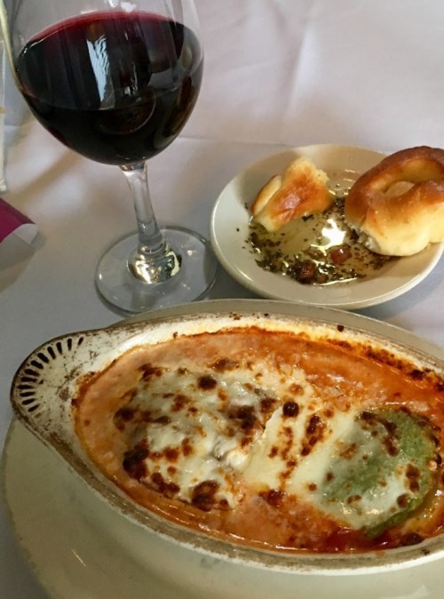 A baked dish with melted cheese, served with a glass of red wine and a plate of bread rolls with olive oil.