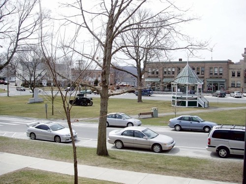 A view of a small town square with a gazebo, parked cars, and bare trees on a cloudy day.