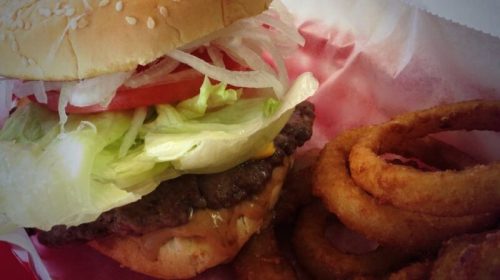 A close-up of a sesame seed bun burger with lettuce, tomato, and onion, served with crispy onion rings.