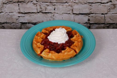 A golden waffle topped with cherry sauce and whipped cream, served on a turquoise plate against a brick background.