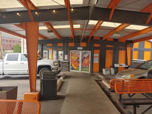 A covered outdoor area with orange and gray walls, featuring a doorway and parked cars. Trash bins and seating are visible.