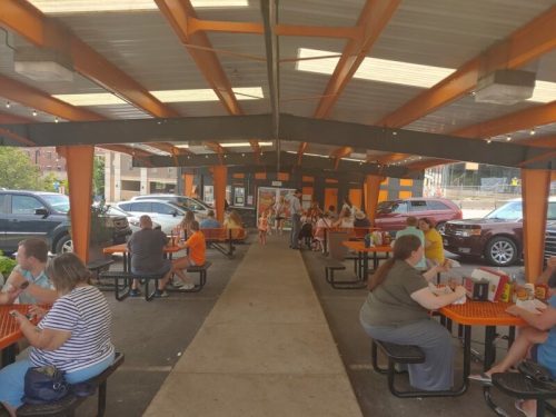 Outdoor dining area with people eating at tables under a covered structure, surrounded by parked cars.