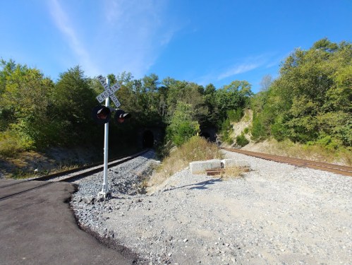A railway crossing sign near two tunnels, surrounded by greenery and a clear blue sky.