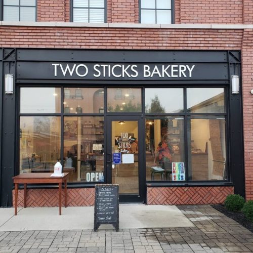 Exterior of Two Sticks Bakery, featuring large windows, a wooden table, and a chalkboard sign outside.