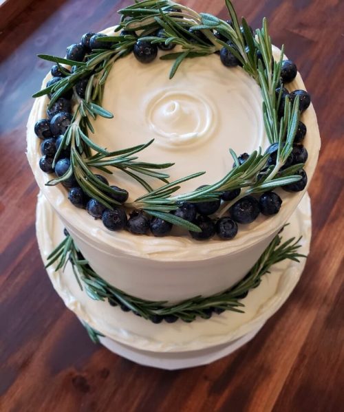 A two-tiered cake decorated with cream frosting, fresh blueberries, and rosemary sprigs on top.