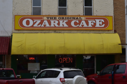 Sign for "The Original Ozark Cafe" with a yellow awning and "OPEN" neon sign in the window. Red and white vehicles parked outside.
