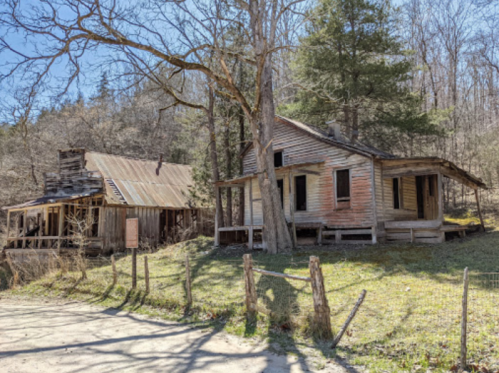 Two old, weathered houses sit on a grassy area surrounded by trees, with a dirt path in the foreground.