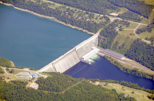 Aerial view of a large dam with water flowing, surrounded by green trees and a calm lake.