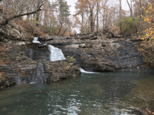 A serene waterfall cascades over rocky terrain into a calm pool, surrounded by autumn foliage and trees.