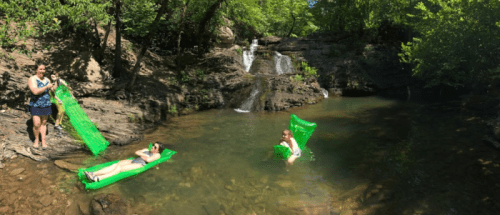 Two people relax on green floaties in a serene pool by a waterfall, surrounded by lush greenery.
