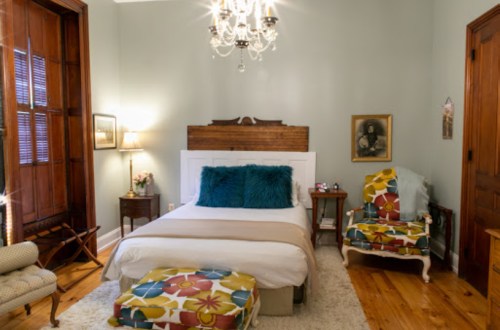 Cozy bedroom with a bed, colorful chairs, a chandelier, and wooden accents, featuring soft lighting and decorative pillows.