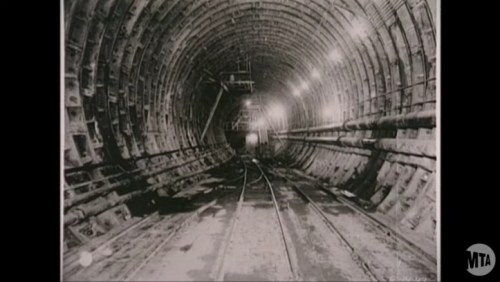 Black and white image of a long, empty subway tunnel with tracks and overhead lights.