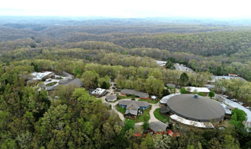 Aerial view of a wooded area with buildings nestled among trees, surrounded by rolling hills and a cloudy sky.