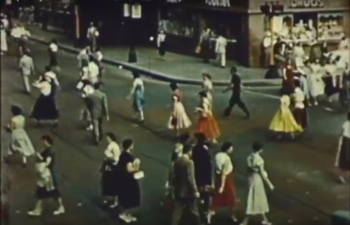A busy street scene from the past, with people in vintage clothing walking along the sidewalk.