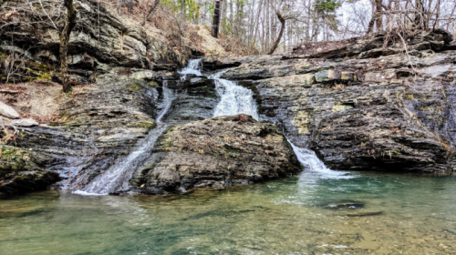 A serene waterfall cascading over rocky terrain into a calm pool, surrounded by trees and natural scenery.