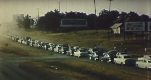 A long line of vintage cars on a highway, with billboards and trees in the background, captured in a hazy atmosphere.