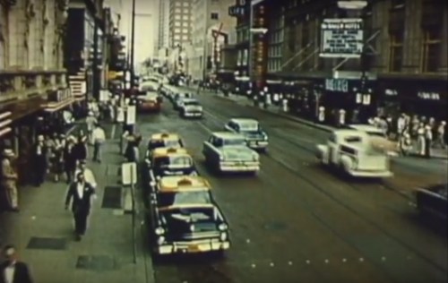 A busy city street in the mid-20th century, featuring taxis, pedestrians, and vintage cars.