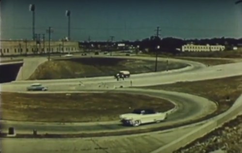 A vintage scene of cars navigating a curved road near a stadium, with buildings and power lines in the background.