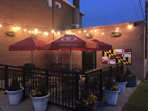 Outdoor patio area with string lights, umbrellas, and potted plants against a brick wall. Maryland flag decor visible.