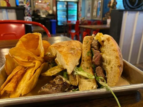 A close-up of a sandwich filled with meat and greens, served with crispy potato chips on a metal tray.