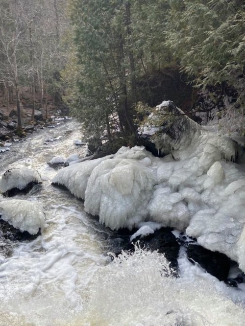 A frozen waterfall cascades over rocks, surrounded by trees in a winter landscape.