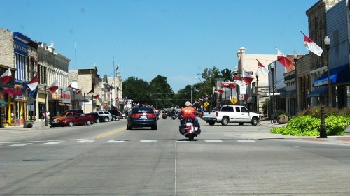A street view of a small town with flags lining the road, featuring cars and a motorcycle in the foreground.