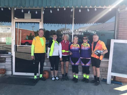 A group of six cyclists in bright gear poses outside the Greenback Drug Store and Diner.