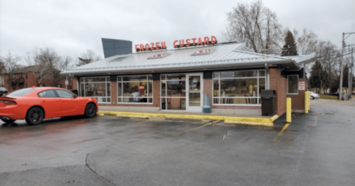 A frozen custard shop with a red car parked outside, featuring a modern design and cloudy sky.