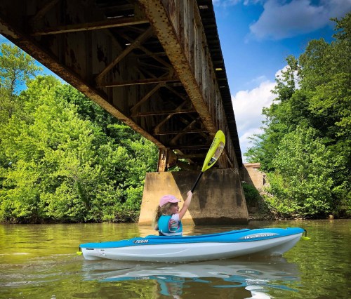 A person in a kayak paddles under a rustic bridge surrounded by lush greenery and a clear blue sky.