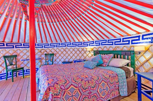 A colorful yurt interior featuring a bed with a patterned blanket and decorative walls, illuminated by soft lighting.