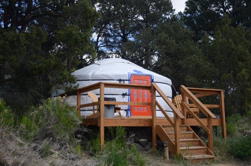 A white yurt with a colorful rug on the porch, surrounded by trees and a wooden deck with stairs.