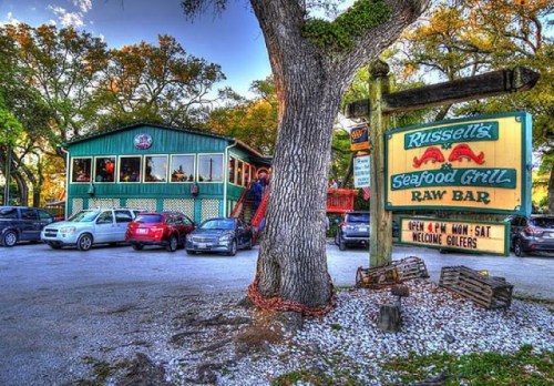 A seafood grill and raw bar with a green building, surrounded by trees and parked cars, welcoming golfers.