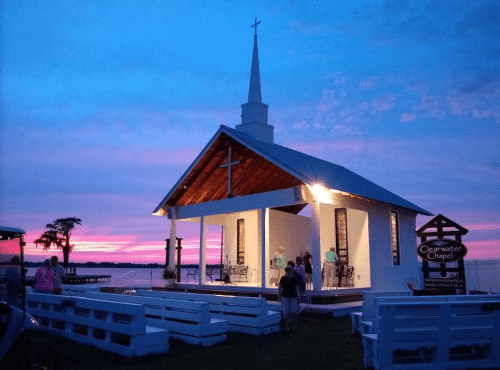 A white chapel with a steeple at sunset, surrounded by palm trees and people, with vibrant pink and purple skies.