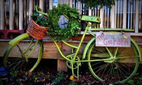 A vintage green bicycle with a basket, decorated with a wreath, next to a sign that reads "The Garden."