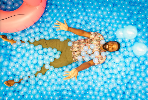 A person lies among blue plastic balls, surrounded by a pink inflatable ring, with arms outstretched and a relaxed expression.