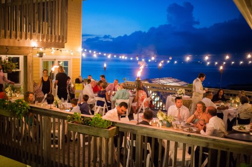A lively outdoor dinner party at dusk, with guests dining under string lights and tiki torches, overlooking the ocean.