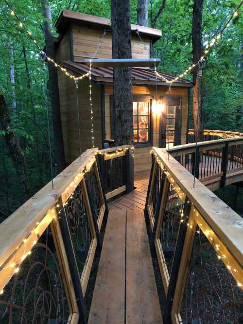 A wooden treehouse connected by a bridge, adorned with string lights, nestled among lush green trees.