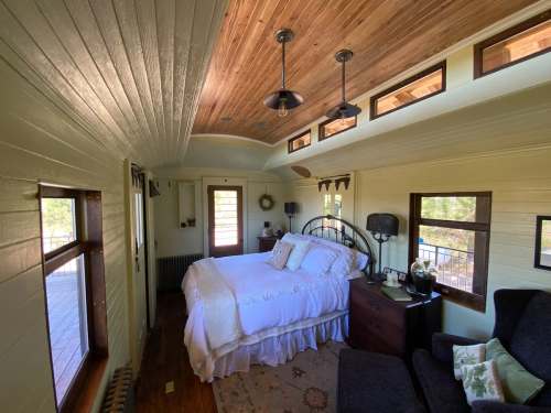 Cozy bedroom with a wooden ceiling, large bed, and natural light from windows, featuring a chair and decorative accents.
