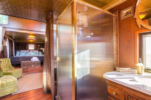 A cozy bathroom featuring a glass shower, wooden vanity, and a view into a stylish bedroom with green armchair.