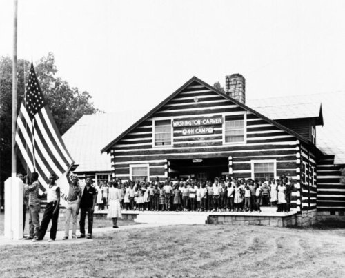 A large group of people stands in front of a log cabin, with a flagpole and American flag in the foreground.