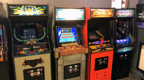 A row of vintage arcade machines, including titles like Star Fox and Smash TV, in a dimly lit game room.
