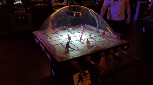 A tabletop hockey game with players under a dome, illuminated in a dim arcade setting.