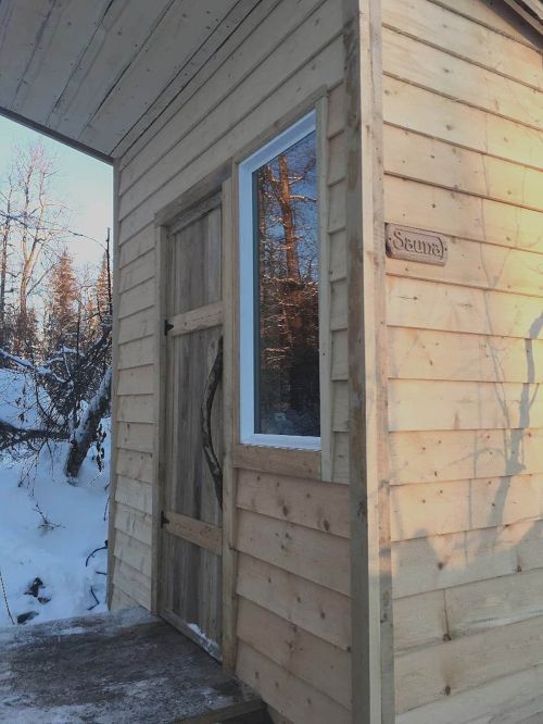 A wooden sauna cabin with a door and window, surrounded by snow-covered trees.