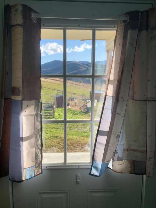 A view through a window with sheer curtains, showcasing rolling hills and a blue sky with clouds.